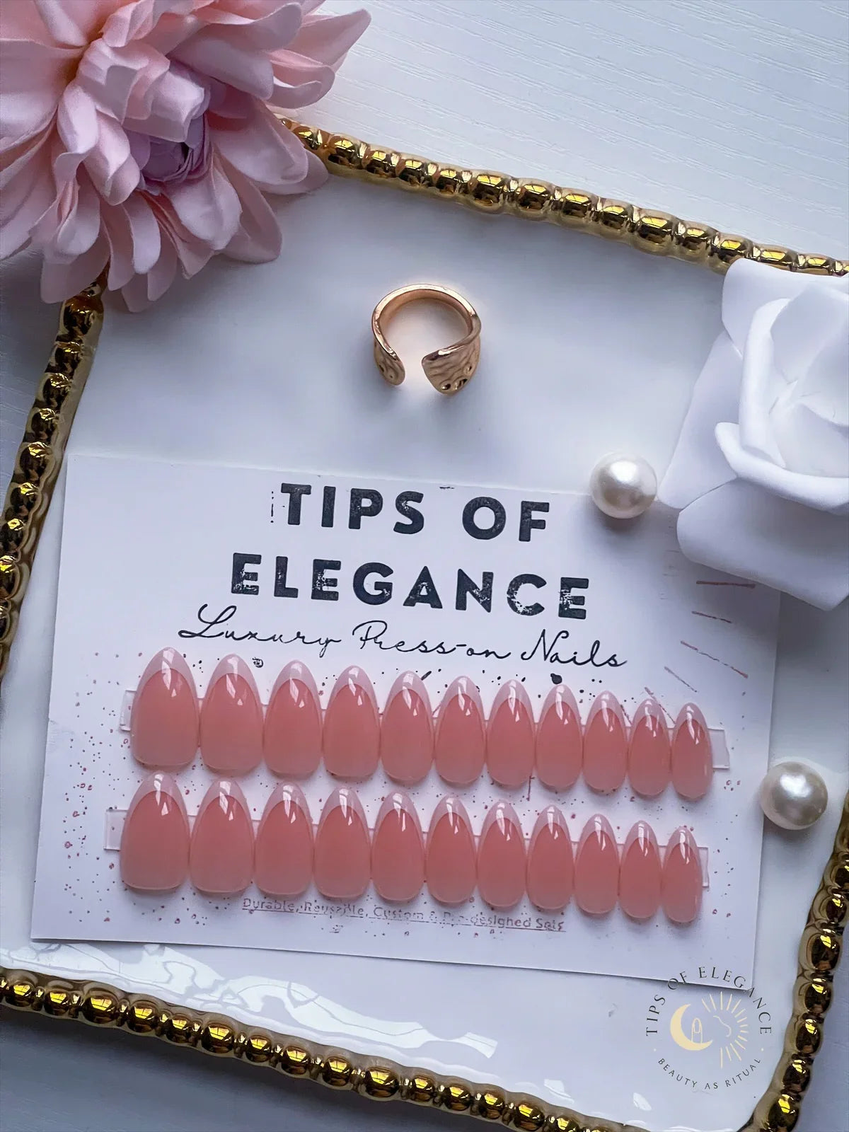 Set of pink false nails on a card labeled 'Tips of Elegance' with a ring and flowers on a decorative tray.