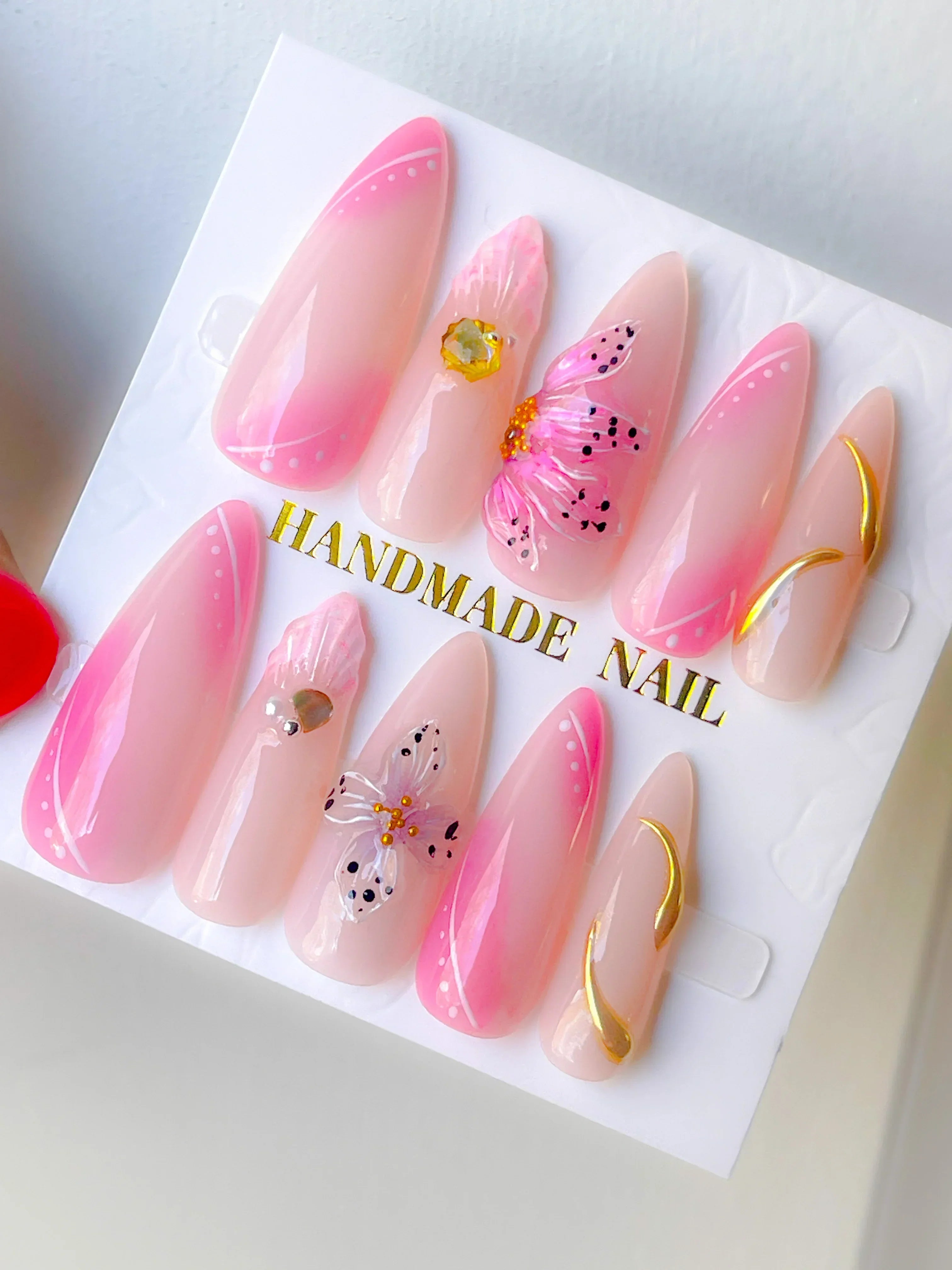 A set of press-on nails with a soft pink ombré design, featuring hand-painted floral accents and gold chrome details.