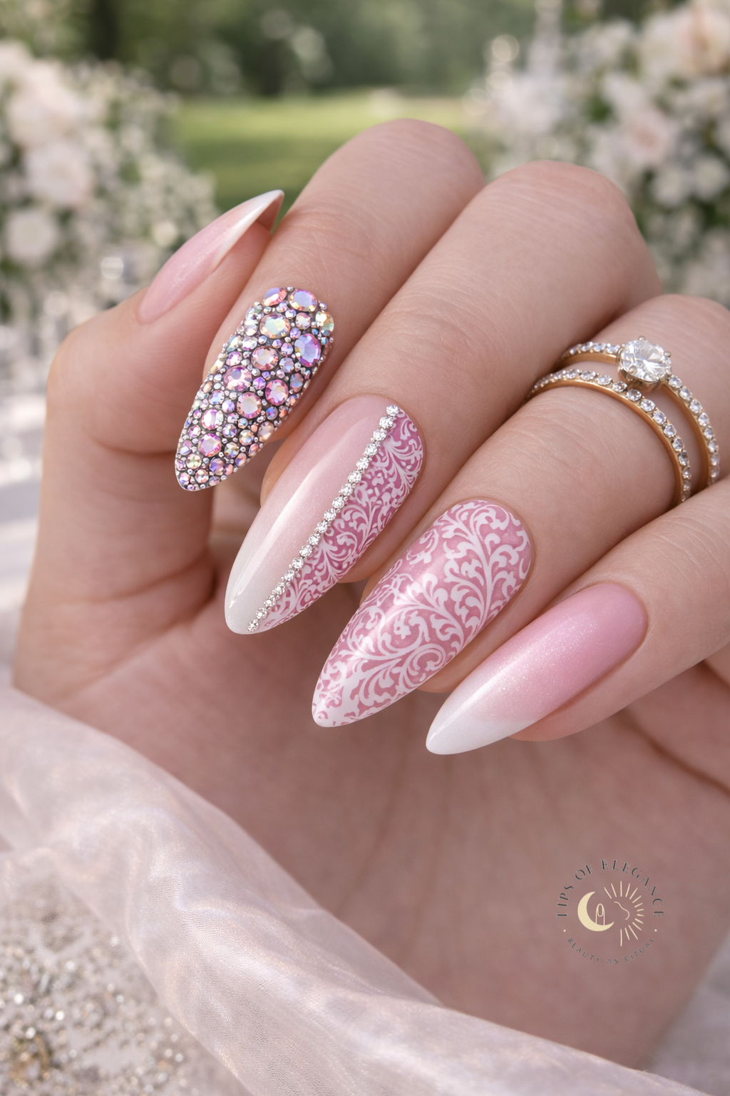 Hand with filigree rolls ab purple and pink crystal gems pink and white baby boomer nails, wearing a ring, against a blurred floral background.